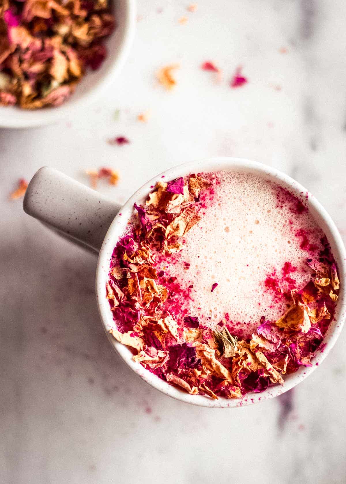 Overhead shot of dairy free rose latte decorated with rose petals. A bowl of petals is nearby.