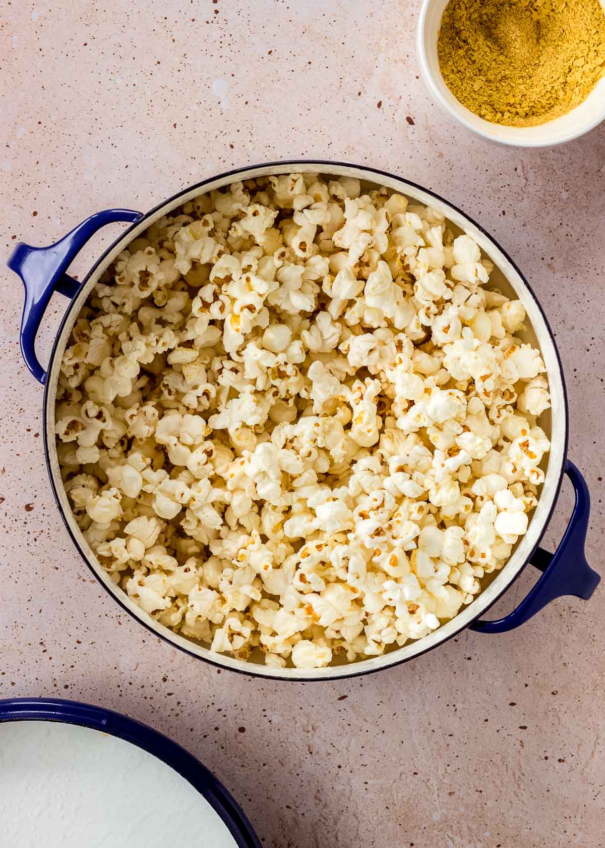 A large blue pot of vegan cheese popcorn with a small bowl of seasoning nearby.