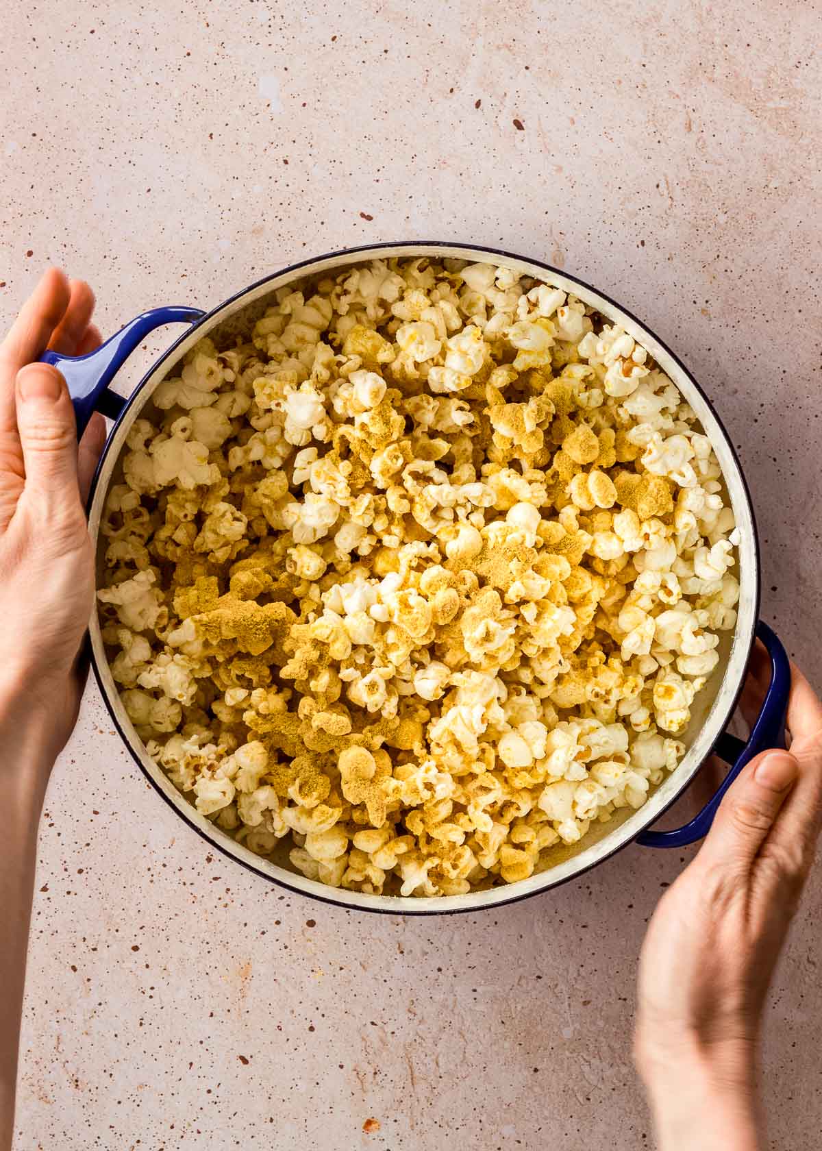 A large blue pot of vegan popcorn with cheesy seasoning being mixed into it. A woman's hands hold the pot handles.