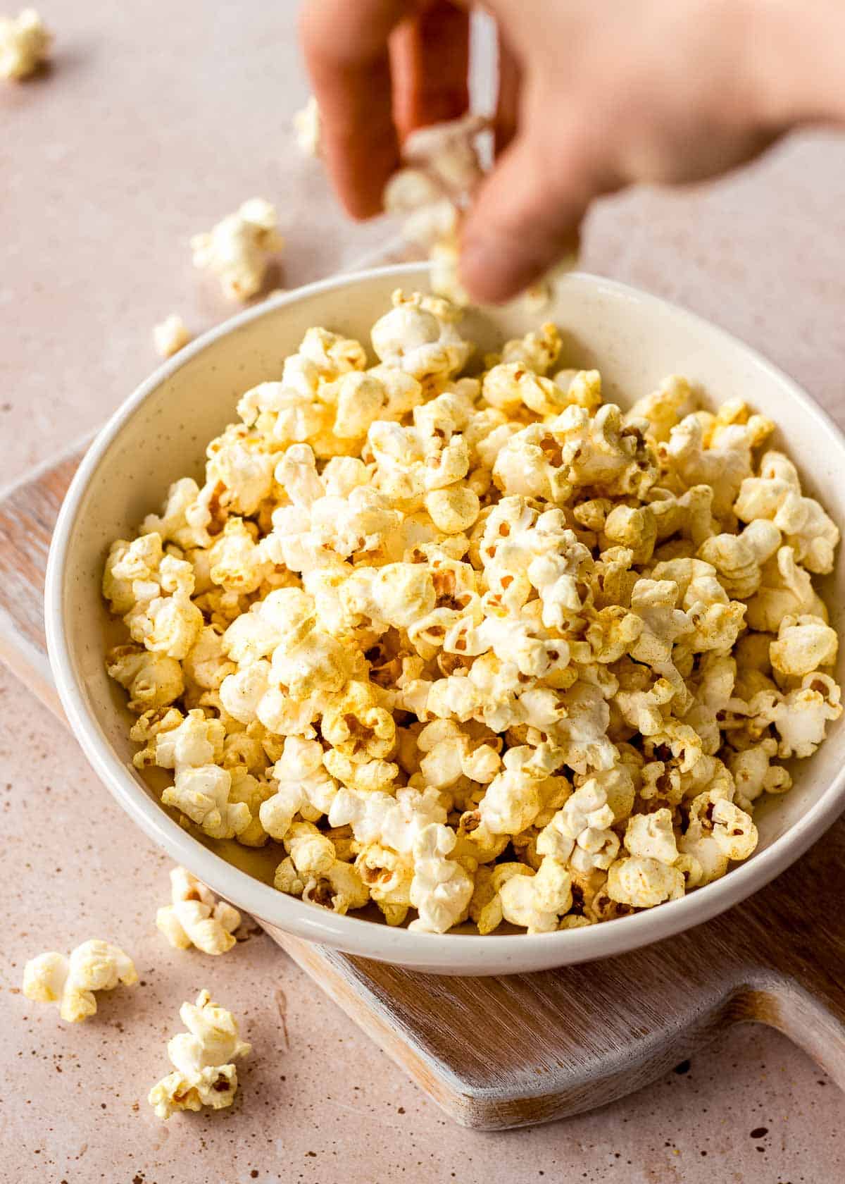 A woman's hand grabs a piece of vegan popcorn from a white bowl on a chopping board. There are pieces of popcorn scattered around.