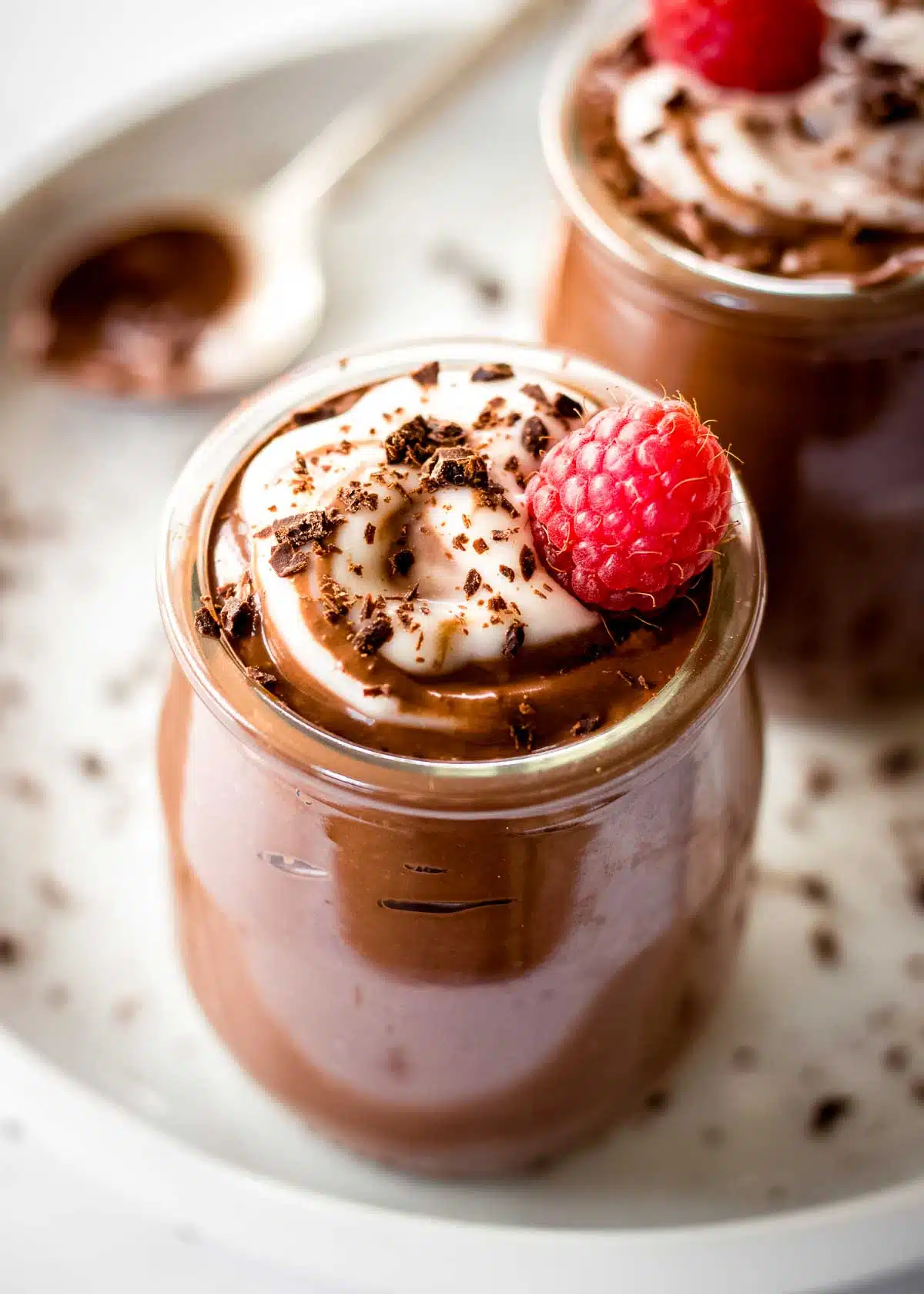 Two glass jars of avocado cacao mousse decorated with cream, shaved chocolate and a raspberry.