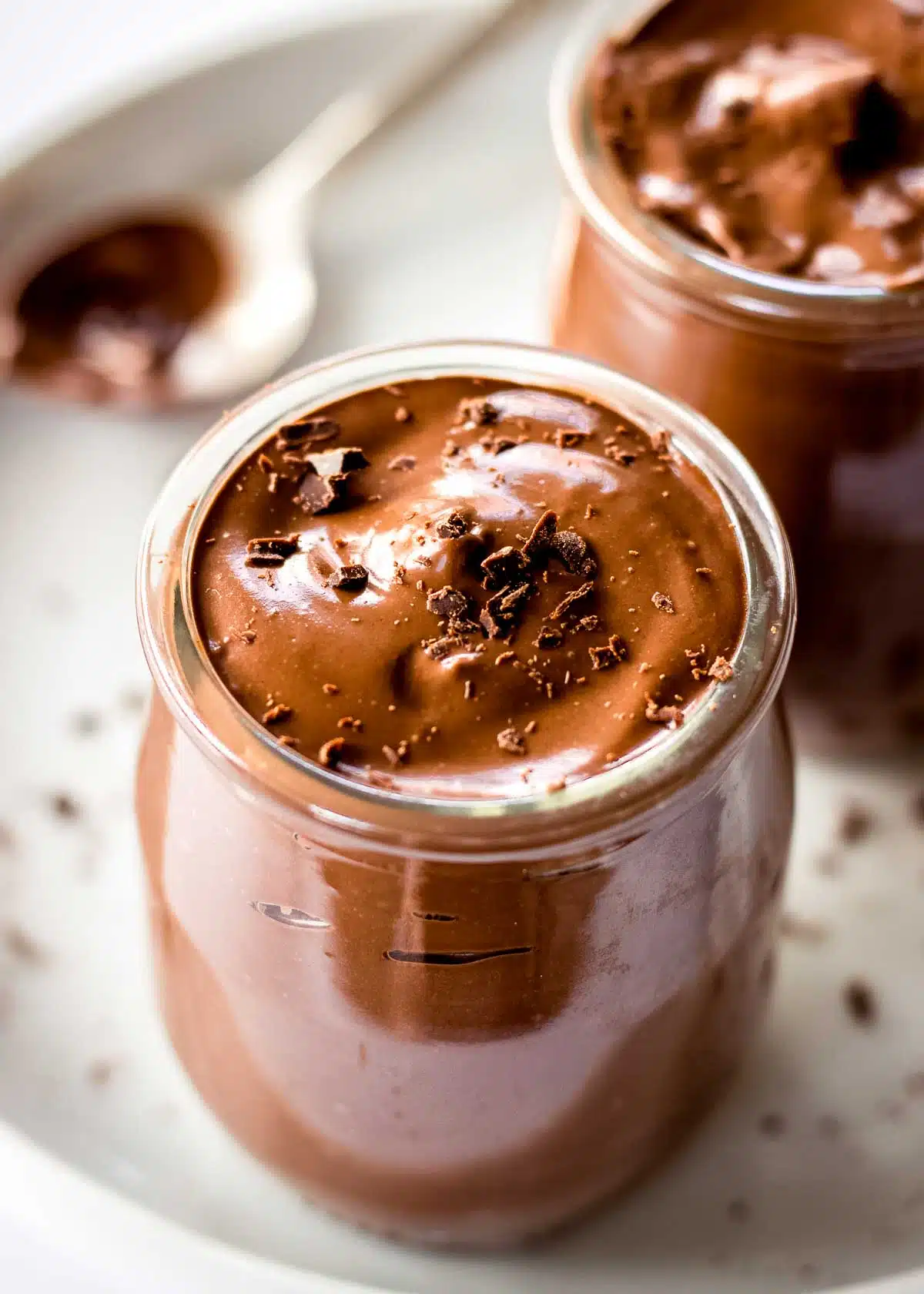 Two glass pots of avocado chocolate mousse topped with grated chocolate. A spoon is off to one side.