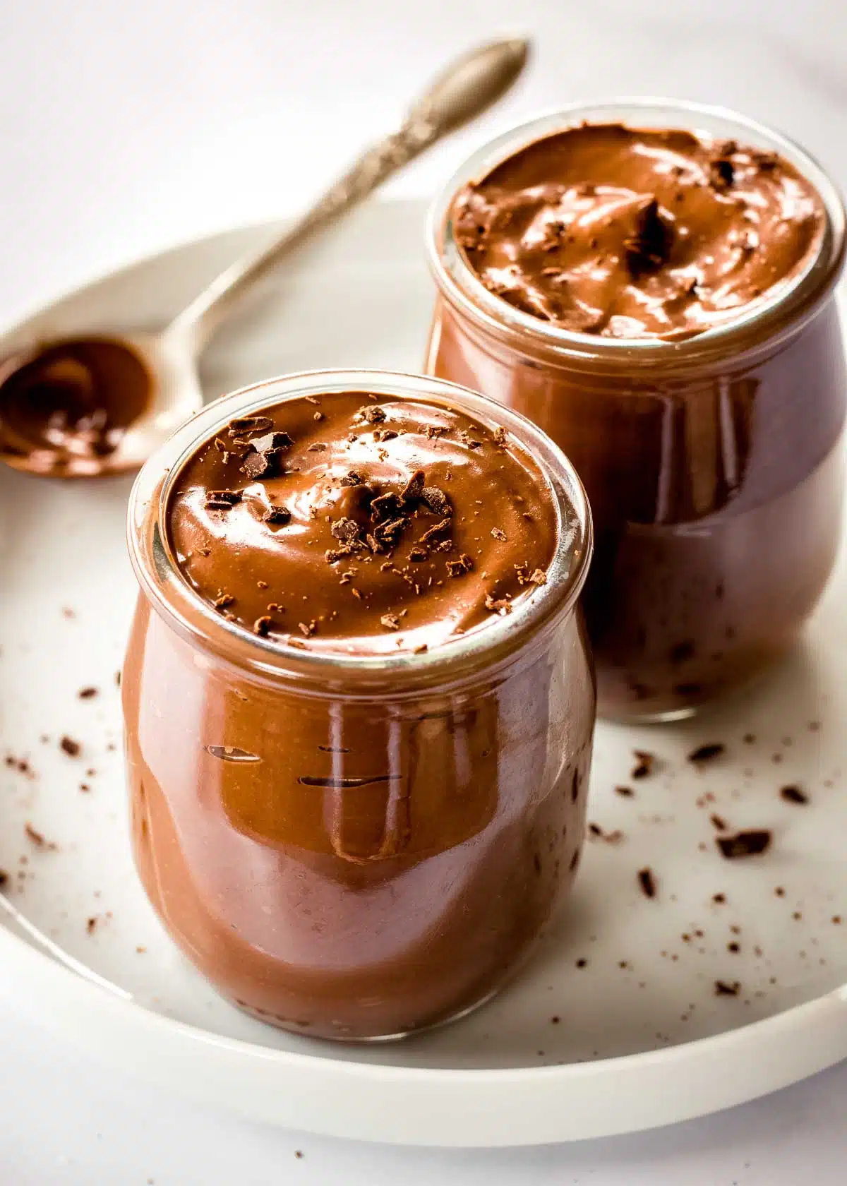 Two glass pots of chocolate avocado mousse on a plate, topped with shaved chocolate. A spoon is off to one side.