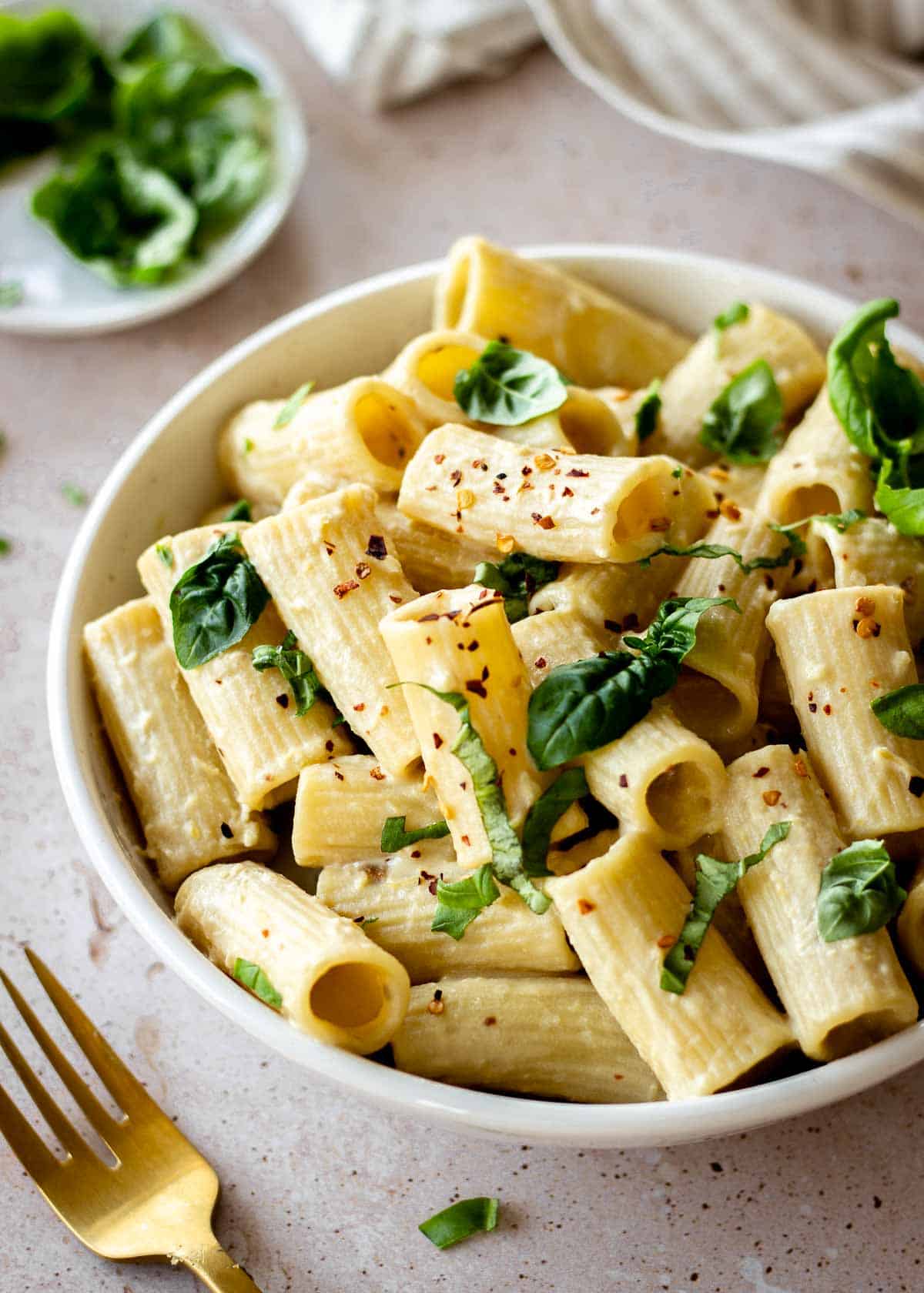 White bowl of pasta with tahini decorated with basil and chili flakes.
