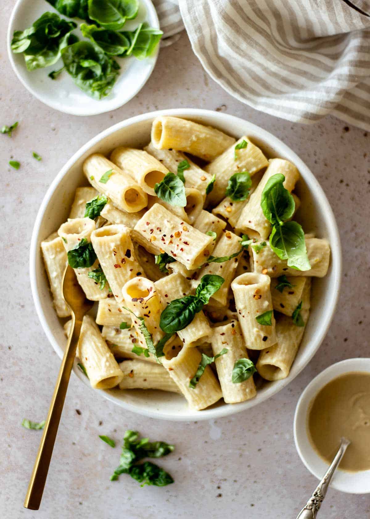 White bowl of tahini pasta decorated with basil and chili flakes. A gold fork sits in the bowl, and a striped napkin and bowls of basil and tahini sit nearby.