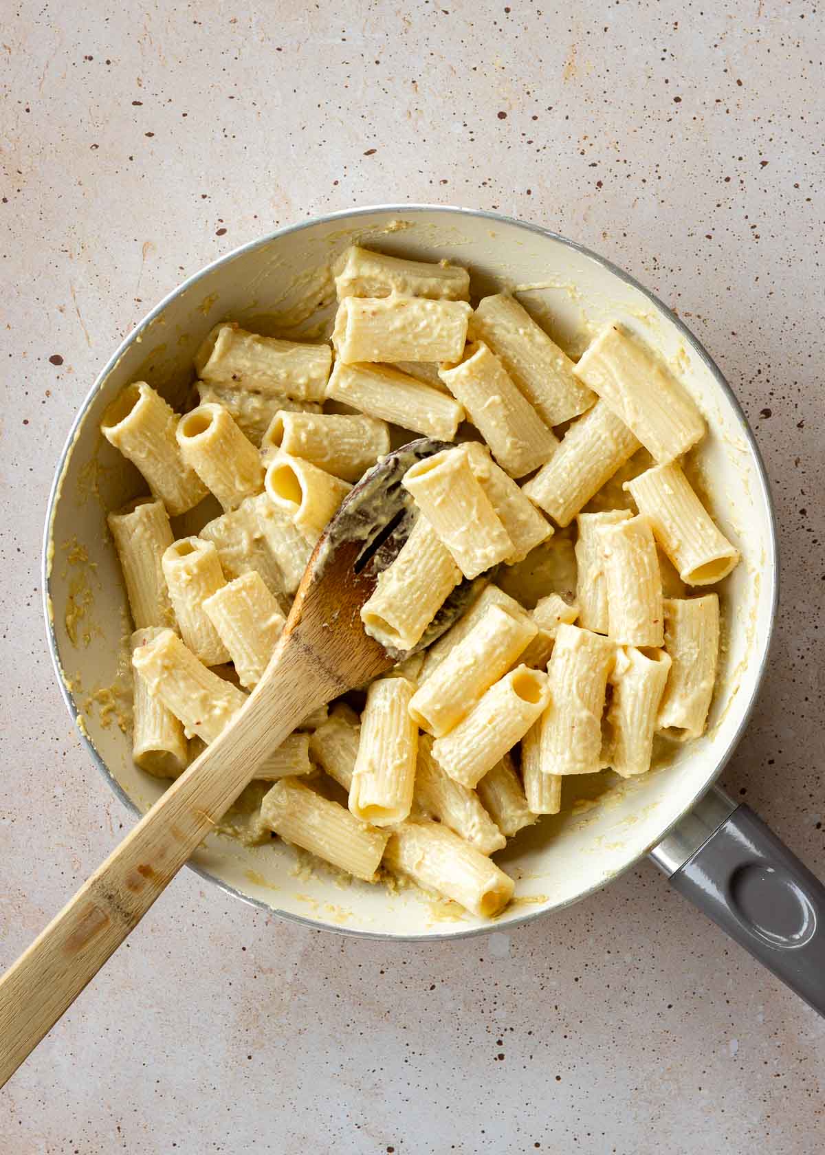 Pasta covered in tahini sauce cooks in a skillet, with a wooden spoon nestled in it.