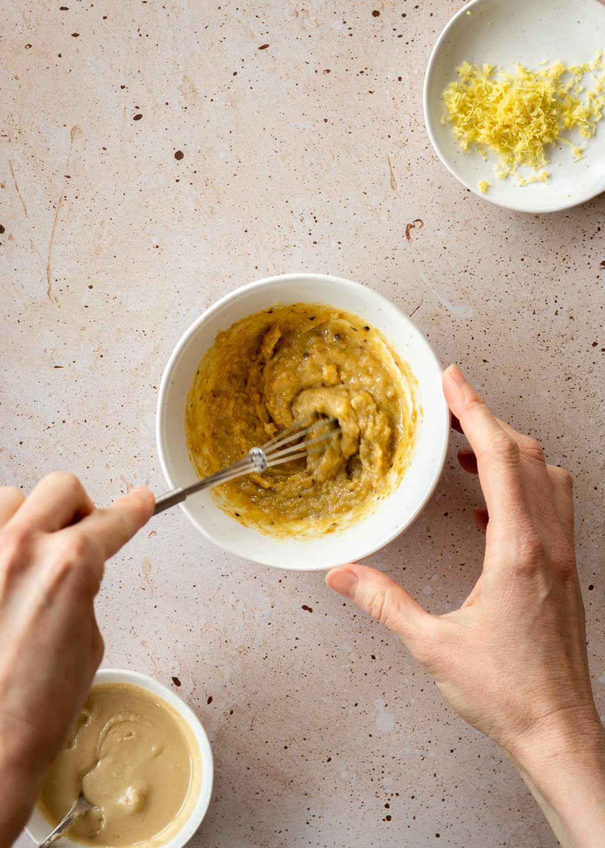 A woman's hands whisk a small bowl of tahini sauce, with dishes of tahini and lemon zest nearby.