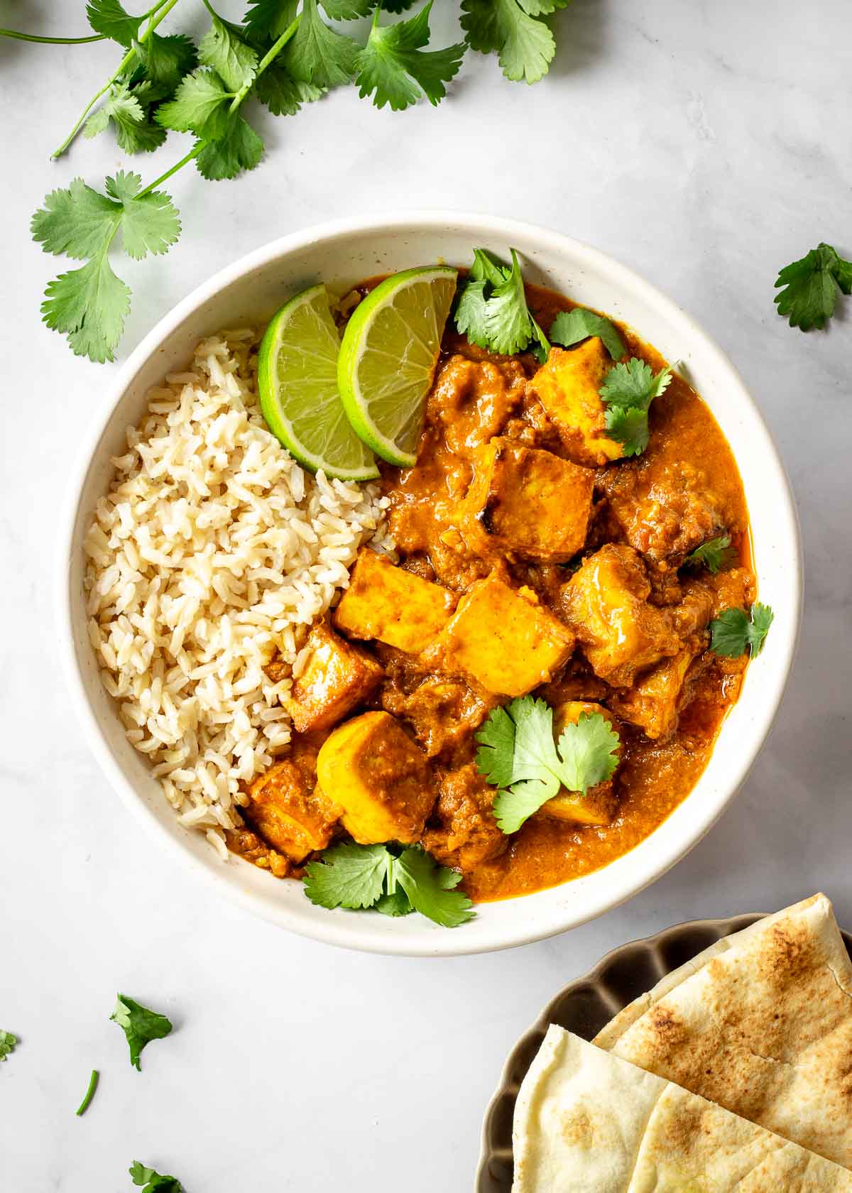 White bowl of tofu butter chicken decorated with cilantro leaves and lime wedges. A dish of naan and some cilantro leaves sit off to one side.