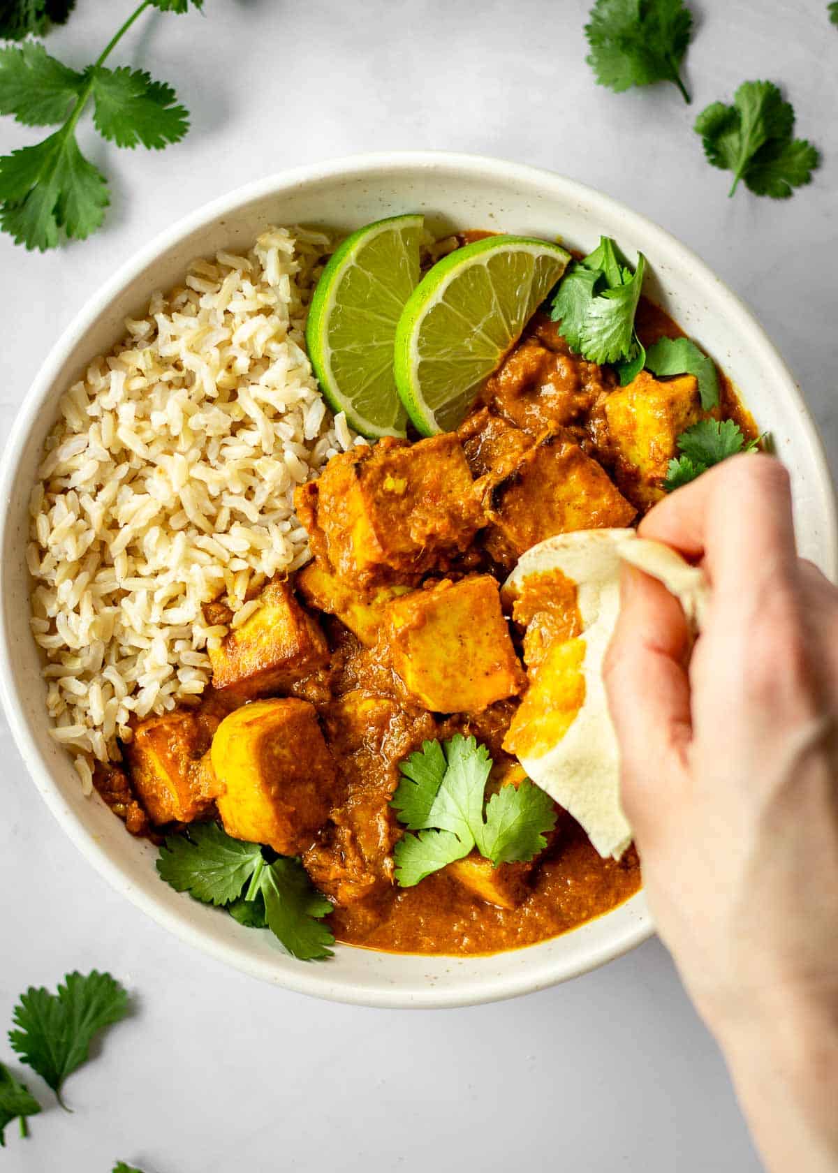 White bowl of vegan butter chicken served with rice, cilantro leaves and lime wedges. A woman's hand scoops up some of it in a piece of naan.