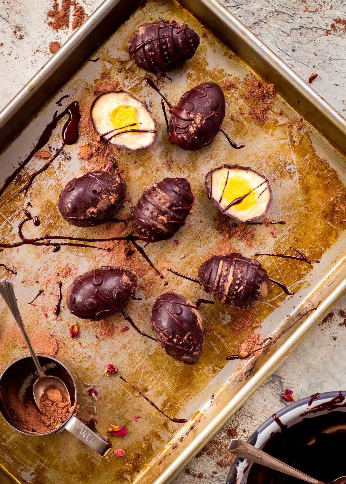 Vegan creme eggs on baking tray drizzled with dark chocolate and with cocoa powder nearby.