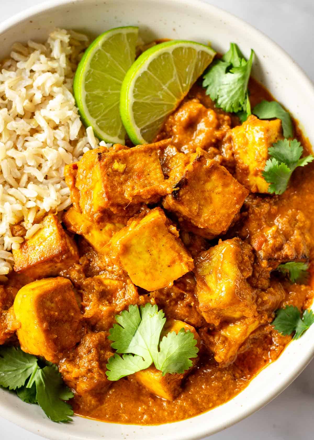 A white dish of vegetarian butter chicken is served with brown rice, lime slices and cilantro leaves.