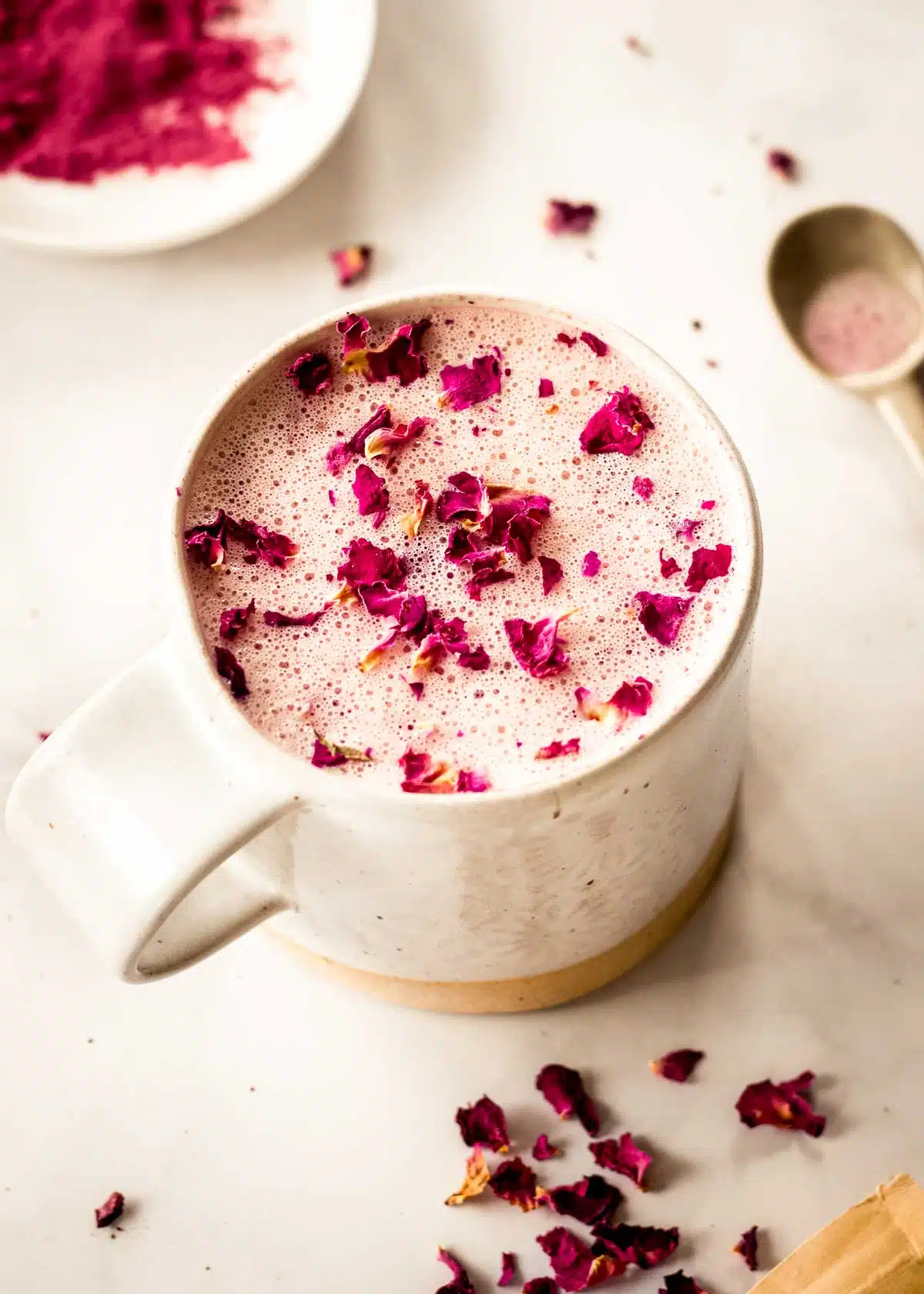A cream coloured mug of pink latte sits on a saucer on a white marble table, decorated with pink rose petals. A dish of beet powder and a spoon sit in the background.