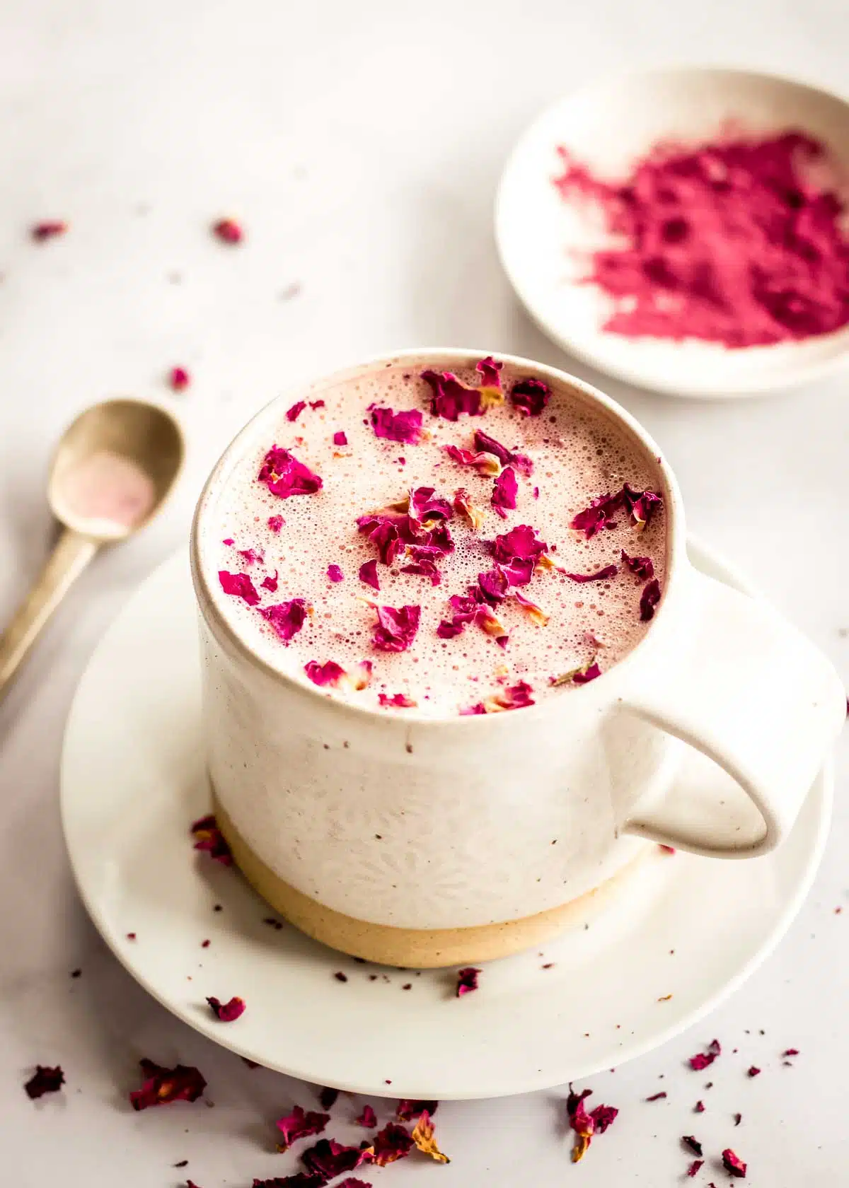 A cream coloured mug of rose latte sits on a saucer on a white marble table, decorated with pink rose petals. A dish of beet powder and a spoon sit in the background.