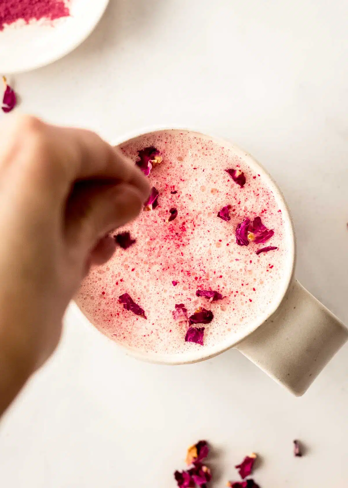 A woman's hand decorates the surface of a rose latte with pink rose petals and beetroot powder.