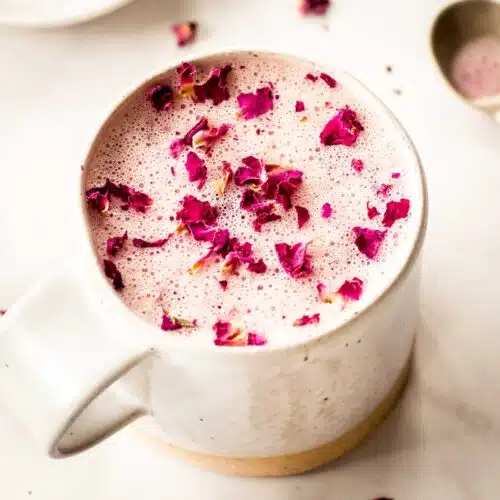 A cream coloured mug of hot rose latte sits on a white marble table, decorated with pink rose petals. A spoon sits nearby.