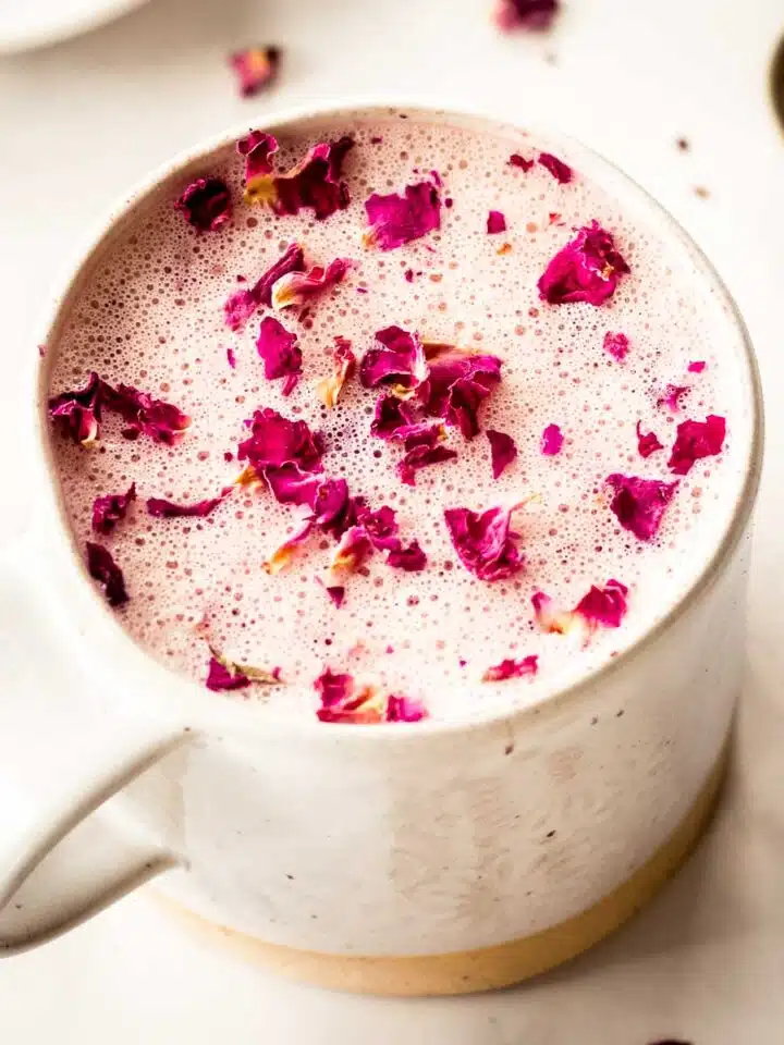 A cream coloured mug of hot rose latte sits on a white marble table, decorated with pink rose petals. A spoon sits nearby.