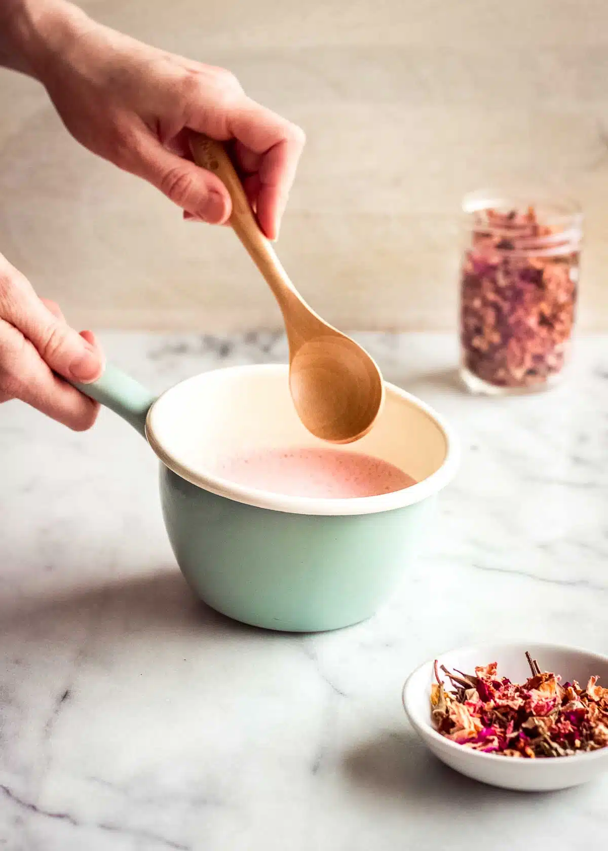 A woman's hand is stirring rose latte ingredients in a turquoise pan, surrounded by containers of rose petals.