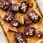 A parchment-lined baking sheet is scattered with vegan snickers bars, which are covered in dark chocolate and decorated with peanuts.
