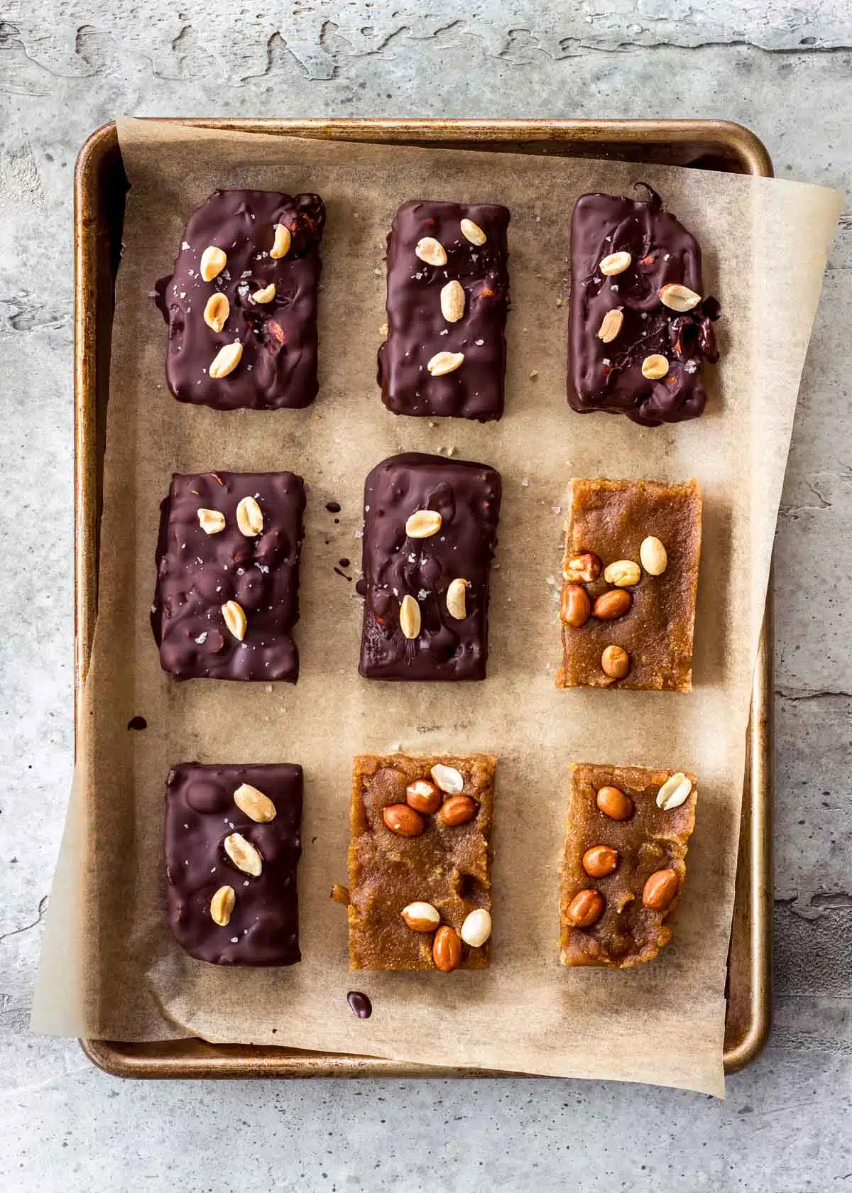 A parchment-lined baking sheet has vegan snickers bars spread over it. Some have already been covered in chocolate.