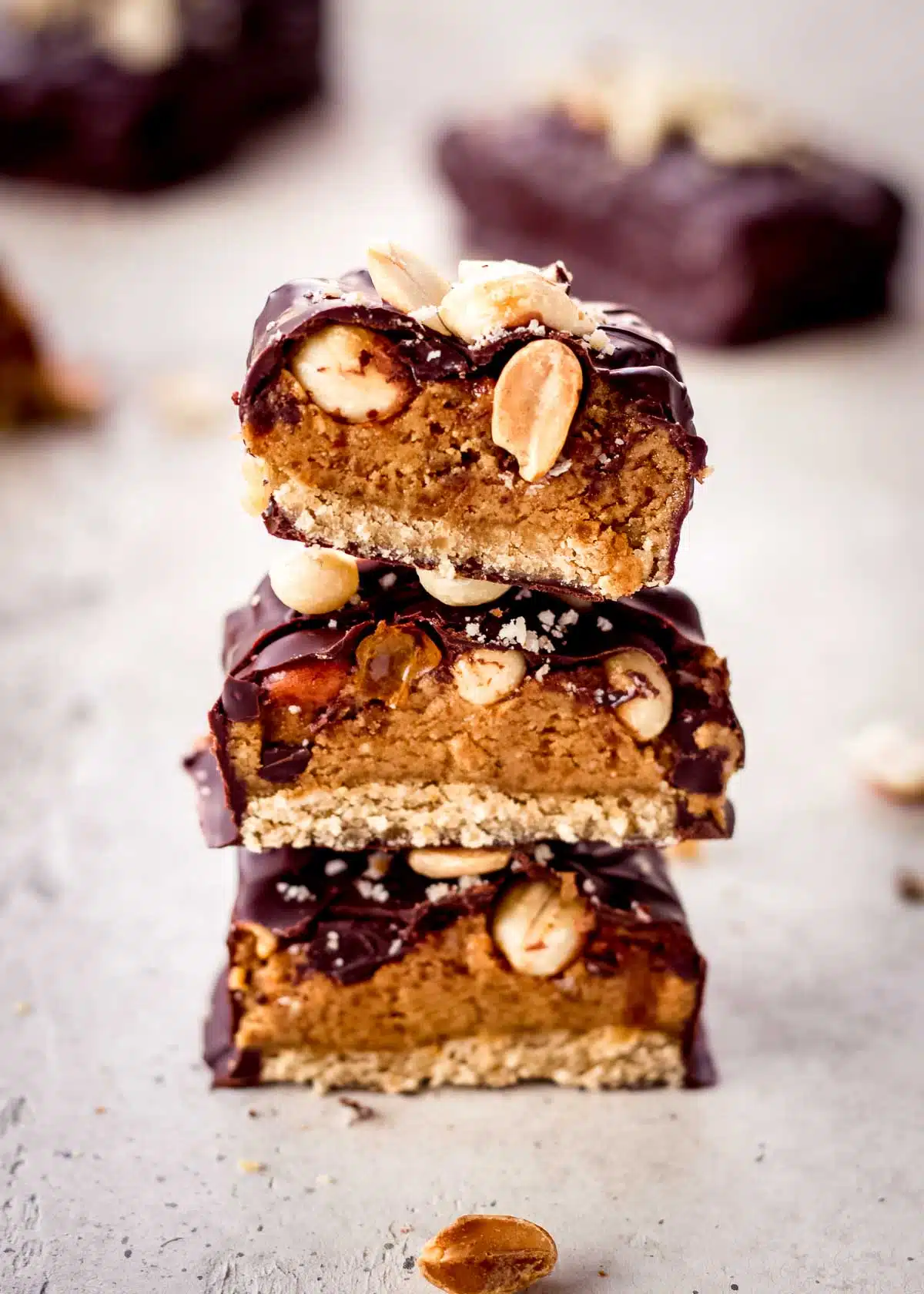 Three homemade vegan snickers bars are stacked together. They are covered in dark chocolate and peanuts are visible.