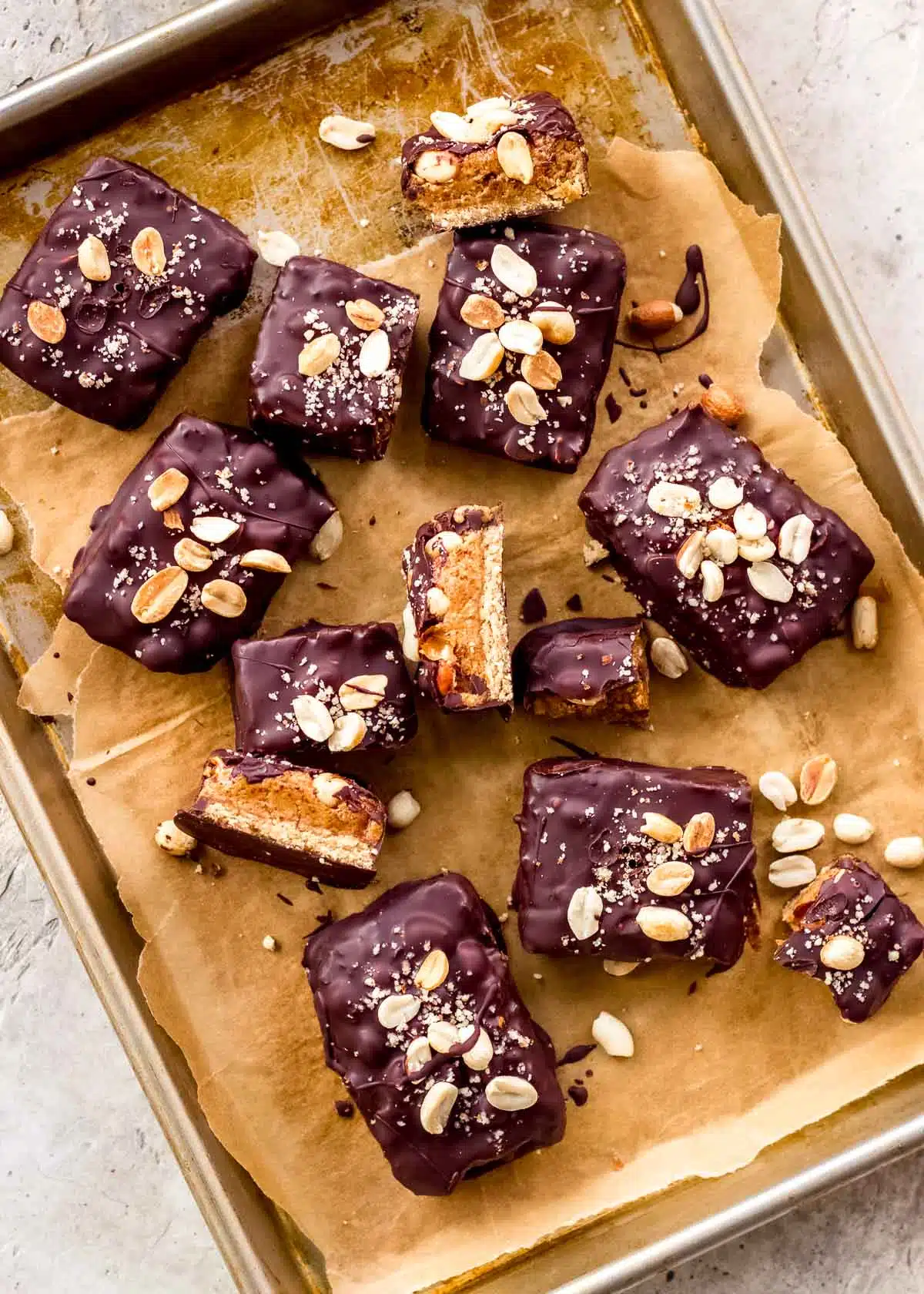 A parchment-lined baking sheet is scattered with vegan snickers bars, which are covered in dark chocolate and decorated with peanuts.