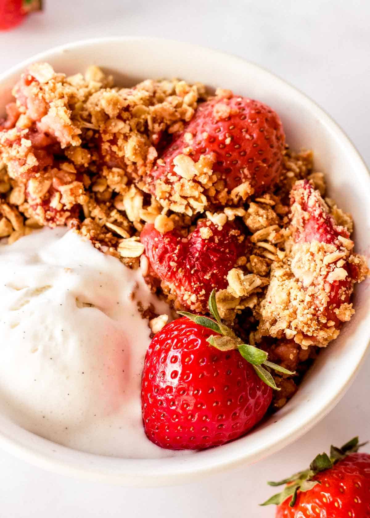 White bowl of strawberry crumble and vanilla ice cream, decorated with strawberries.