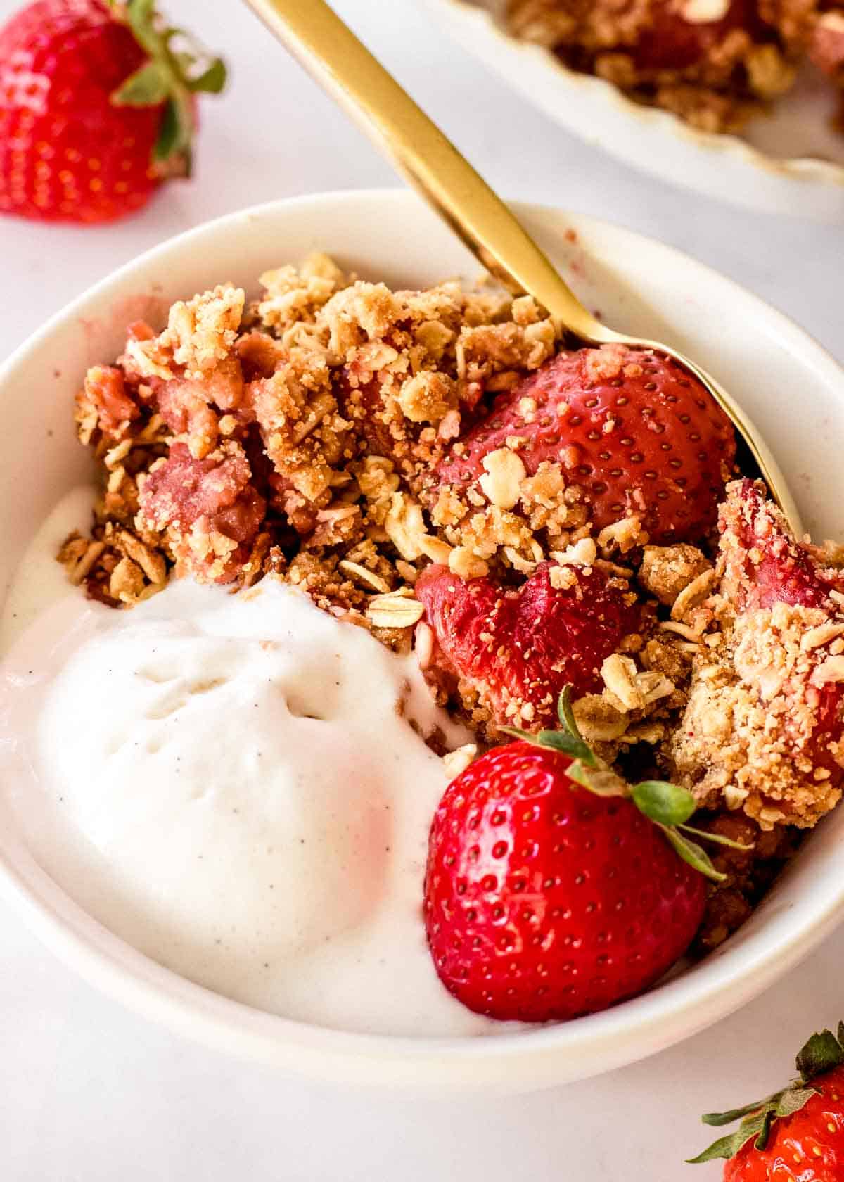 White bowl of strawberry crumble and vanilla ice cream, decorated with strawberries and a golden spoon.