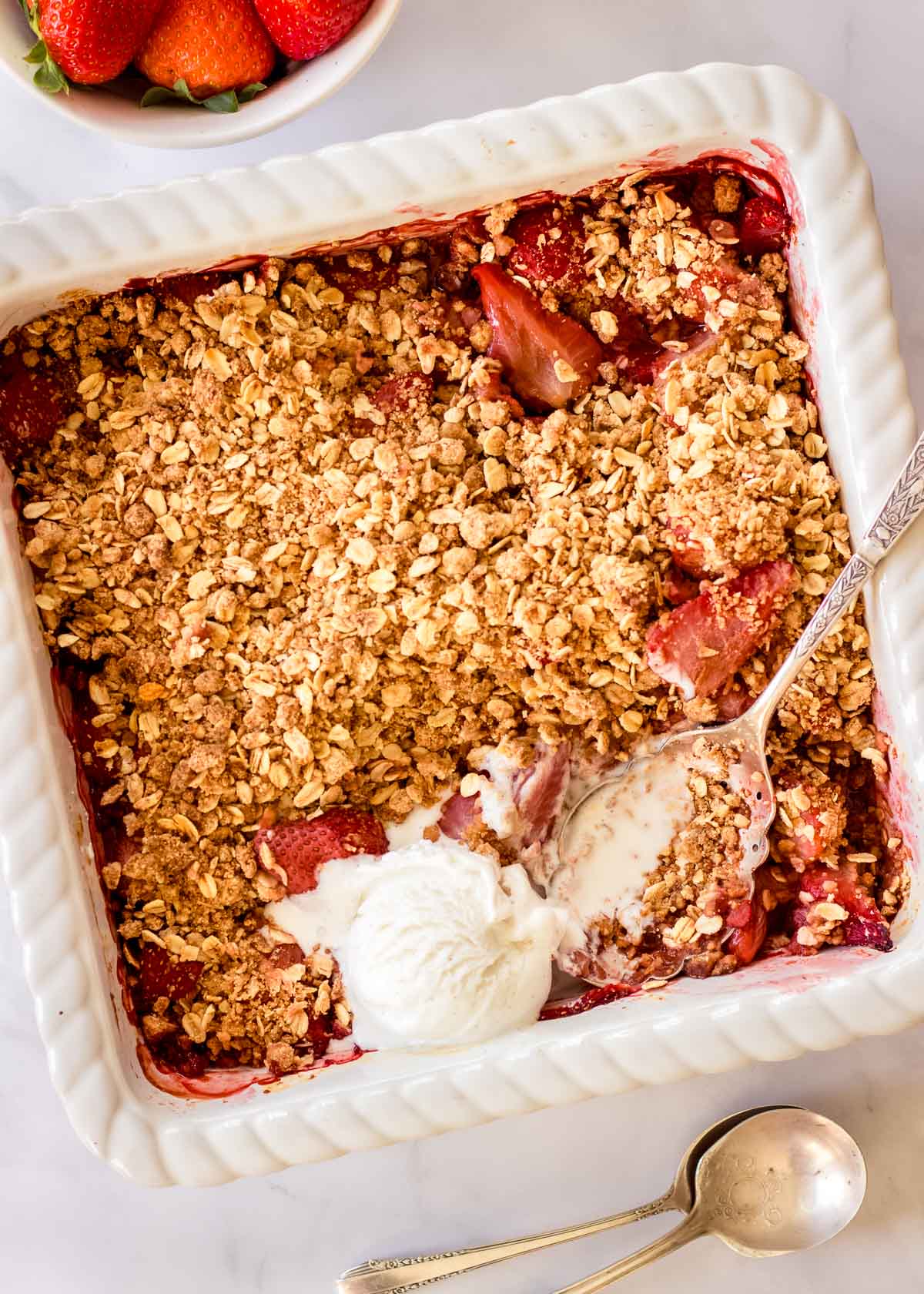 Large white ovenproof dish contains vegan strawberry crumble with oat topping and a scoop of vanilla ice cream. A large fluted silver spoon sits in it.