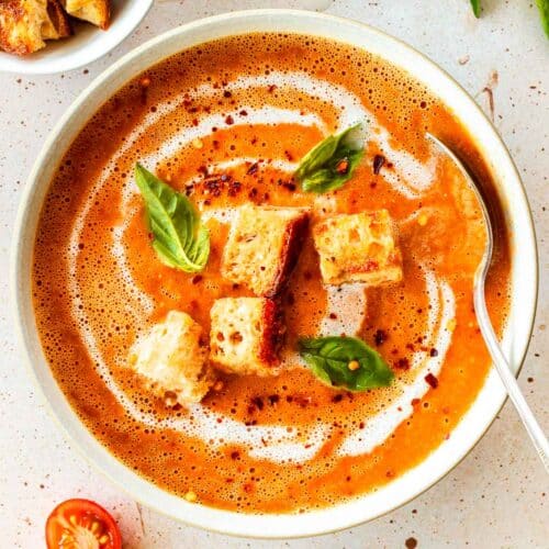 A white bowl of cherry tomato soup decorated with croutons, basil and red pepper flakes.