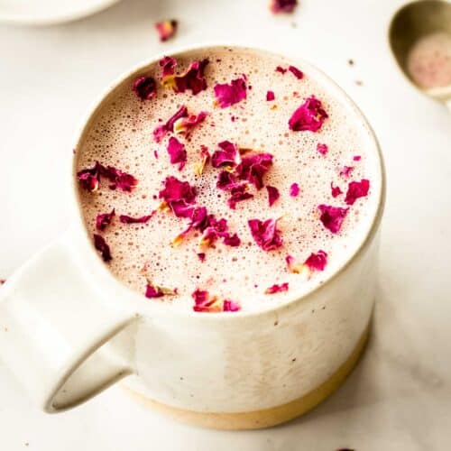 A cream coloured mug of hot or iced rose latte sits on a white marble table, decorated with pink rose petals. A spoon sits nearby.