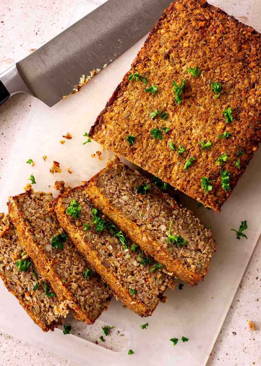 Slices of vegan nut roast on a white chopping board decorated with parsley, with a knife nearby.