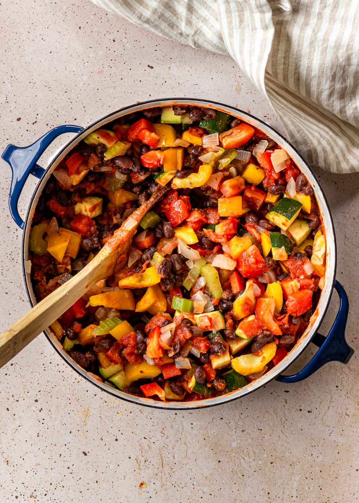 Partially cooked dark blue pot of vegan black bean chili with a spoon in, showing multi-coloured vegetables and black beans, with a striped napkin nearby.