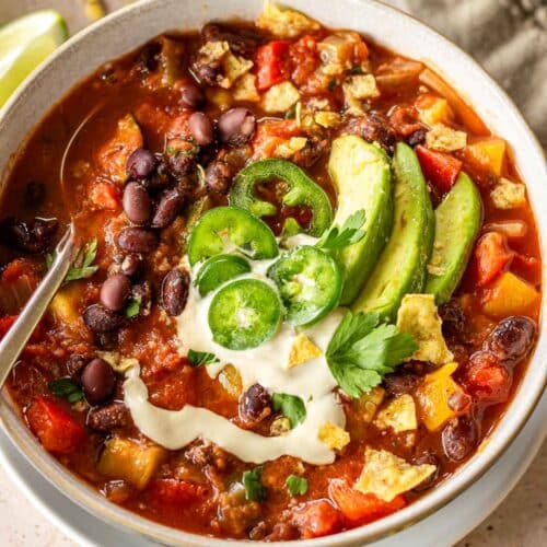 White bowl of vegan black bean chili decorated with avocado slices, jalapeños, coriander and sour cream. Broken tortilla chips are visible on the chili.