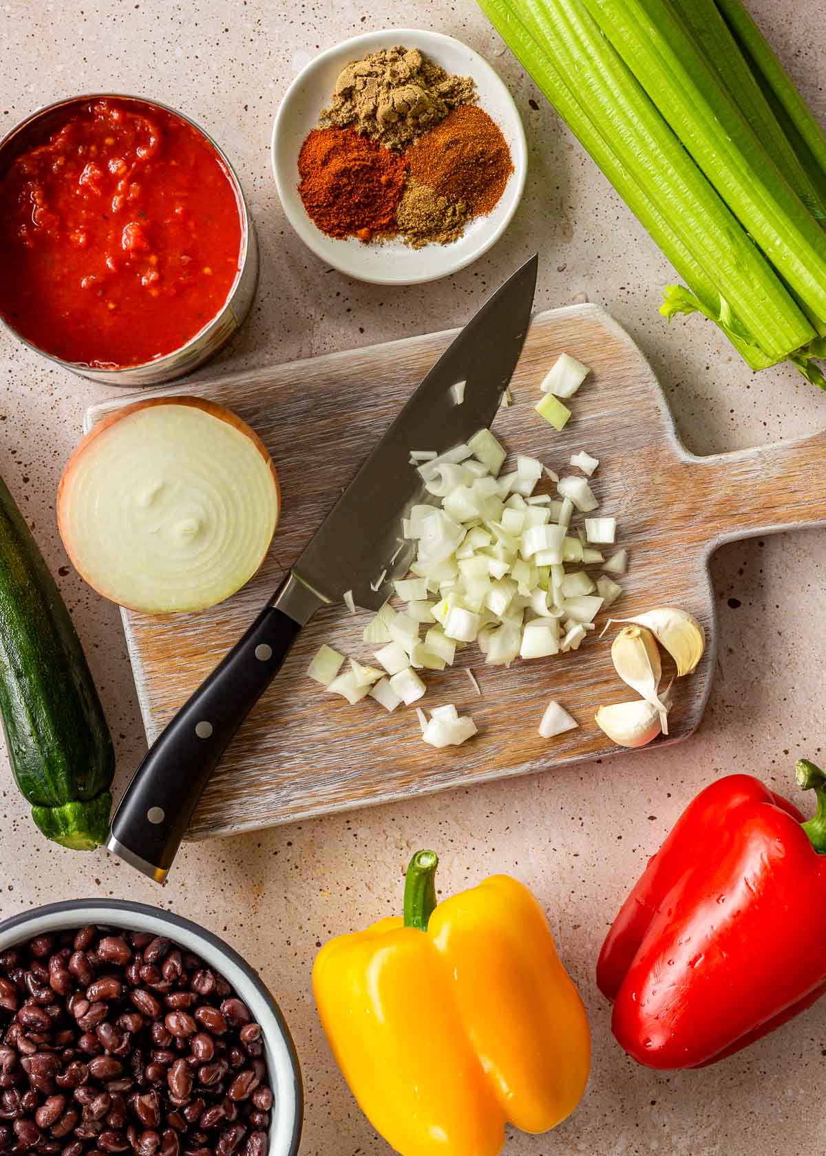 Ingredients for vegan black bean chili, including an onion being sliced on a chopping board, celery, red and yellow bell peppers and a bowl of black beans.