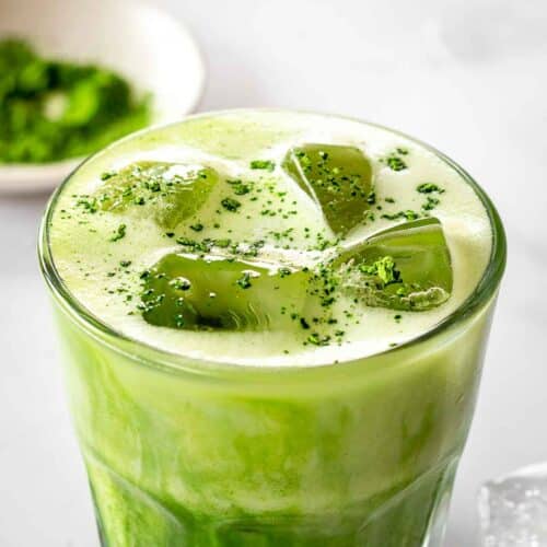 A glass of bright green iced matcha latte with sprinkles of matcha powder and visible ice cubes on top. A bowl of green matcha powder sits nearby.