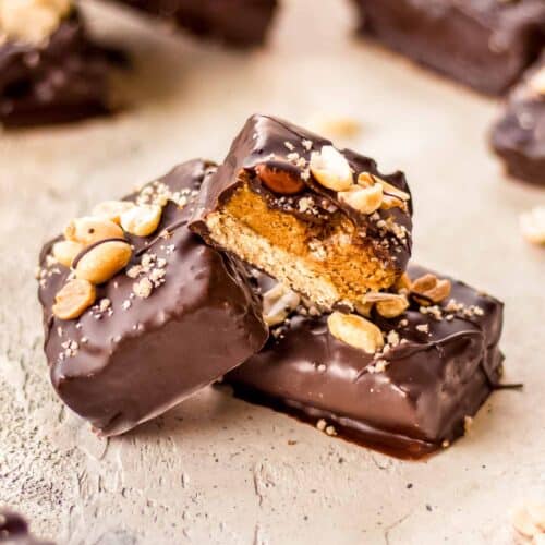 A cluster of three vegan snickers bars sit on a grey surface. One of them has been cut in half to show its layers of oat base, date caramel and chocolate.